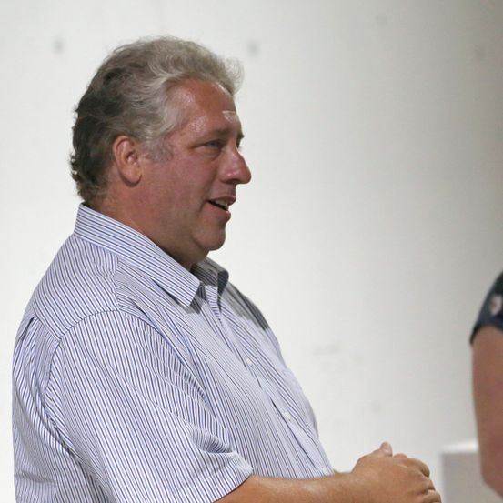 Ein Mann mit grauem Haar, der ein blau-weiß gestreiftes Hemd trägt, lächelt und schaut nach rechts.