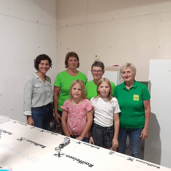 Fünf Frauen und zwei Mädchen stehen in einem Raum und lächeln für ein Foto. Die Frauen tragen grüne Shirts und die Mädchen tragen rosa und weiße Shirts. Ein Tisch mit einem weißen Tuch hat das Wort 'Postleitzone' darauf.