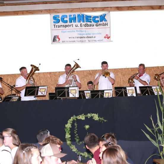 Eine Blechbläsergruppe spielt auf der Bühne, über ihnen hängt ein Banner mit der Aufschrift 'Schneck Transport u. Erdbau GmbH'. Das Publikum sitzt vor ihnen.