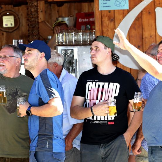 Eine Gruppe von Männern in einer Bar, einer trägt ein schwarzes T-Shirt mit weißen Texten und hält ein Bierglas.