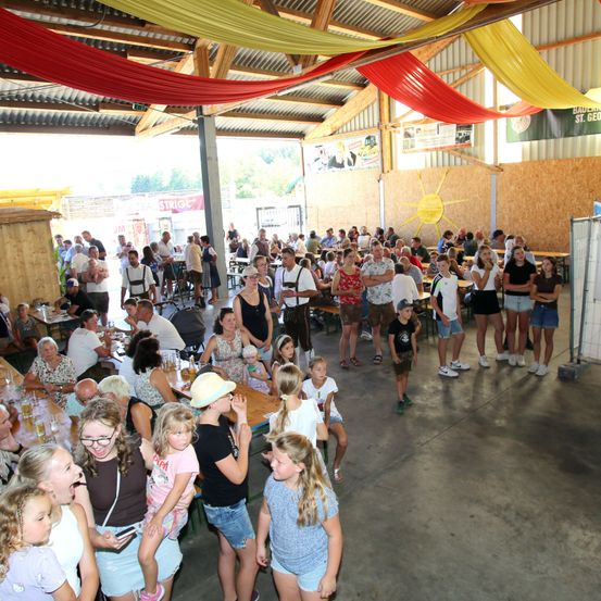 Eine große Gruppe von Menschen versammelt sich in einem Saal, wahrscheinlich für ein Festival. Einige sitzen an Tischen, während andere herumstehen. Der Saal ist mit bunten Bannern und einem Sonnendesign an der Wand geschmückt.
