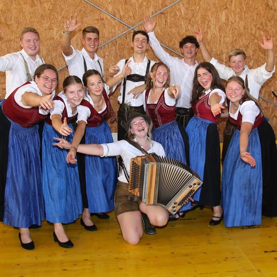 Eine Gruppe junger Menschen in traditioneller deutscher Kleidung posiert für ein Foto, wobei eine Person ein Akkordeon spielt.