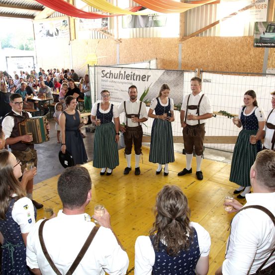 Eine Gruppe von Menschen in traditioneller bayerischer Kleidung führt auf einer Bühne auf, hinter ihnen ein Banner. Einige halten Getränke in den Händen, andere schauen zu.