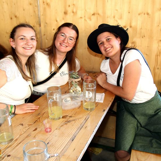 Drei Frauen sitzen an einem Holztisch, lächeln und posieren für ein Foto. Sie tragen Freizeitkleidung und haben Getränke vor sich. Die Frau rechts trägt einen Hut.