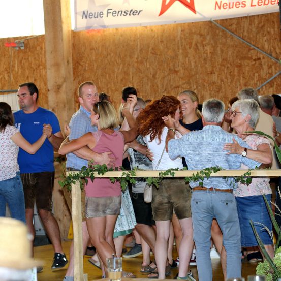 Eine Gruppe von Menschen tanzt in einem Raum mit einer Holzwand und einem Banner mit dem Text 'Neue Fenster'. Einige tragen Hüte und Brillen, andere tragen Sandalen.