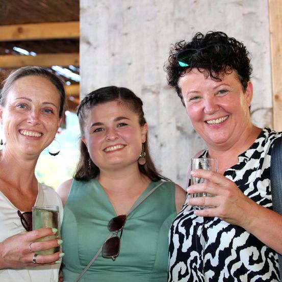 Drei Frauen stehen zusammen, lächeln und halten Gläser mit Wasser. Die Frau auf der linken Seite trägt ein weißes Oberteil und Ohrringe. Die Frau in der Mitte trägt ein grünes Kleid und hat eine Sonnenbrille auf der Schulter. Die Frau auf der rechten Seite trägt ein schwarz-weiß gemustertes Oberteil und hat eine Sonnenbrille auf dem Kopf.