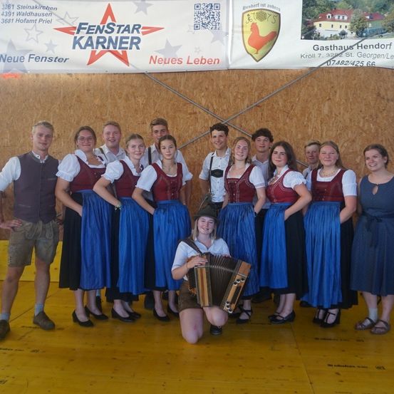Eine Gruppe von Menschen in traditioneller Kleidung, darunter eine Frau mit einem Akkordeon, posiert für ein Foto auf der Bühne.