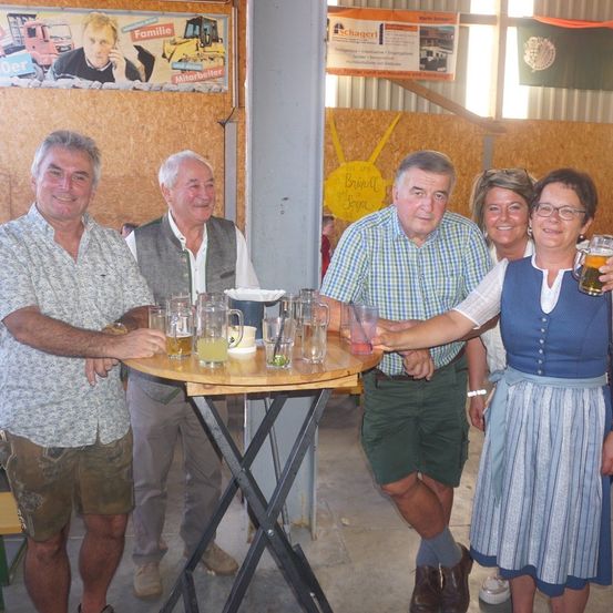Eine Gruppe von Menschen steht um einen Tisch mit Getränken und posiert für ein Foto. Der Tisch hat Gläser, eine Karaffe und Tassen. Im Hintergrund ist eine Wand mit Plakaten und einer Flagge zu sehen.