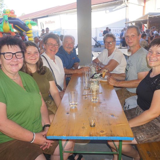 Eine Gruppe von Menschen sitzt an einem Tisch mit Getränken und Essen, lächelt und posiert für ein Foto. Dahinter steht ein Gebäude mit einem Banner und einem Hüpfburg.