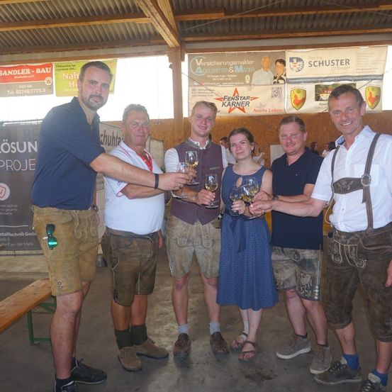 Eine Gruppe von Menschen in traditioneller deutscher Kleidung hält Weinglaser in einem Raum mit Bannern im Hintergrund.