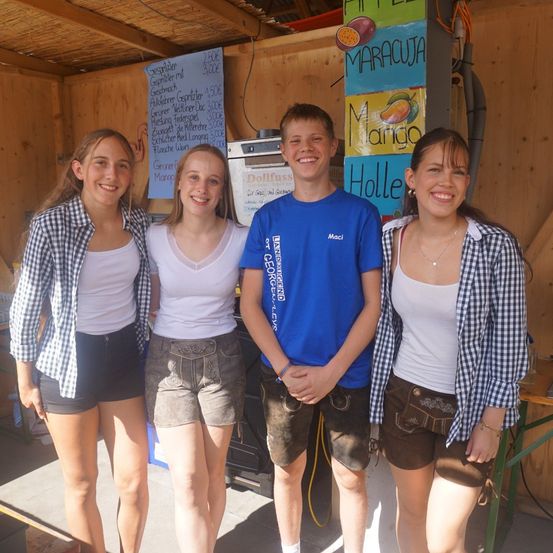 Vier junge Menschen, zwei Mädchen und zwei Jungen, stehen zusammen in einem Raum. Sie alle lächeln und scheinen für ein Foto zu posieren. Hinter ihnen sind Holzwände mit Postern und einem Schild.