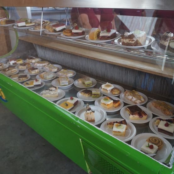 Eine Vitrine voller verschiedener Kuchen und Gebäcke, jeweils auf einem weißen Teller, in Reihen angeordnet. Die Vitrine ist grün mit einer hölzernen Oberseite.