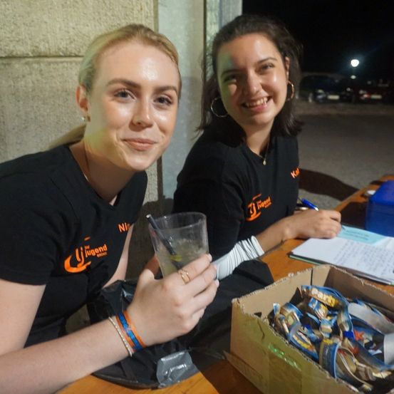 Zwei Frauen in schwarzen T-Shirts mit orangefarbenen Logos sitzen an einem Tisch, eine hält ein Getränk, die andere einen Stift und Papier. Eine Schachtel mit Gegenständen steht auf dem Tisch.