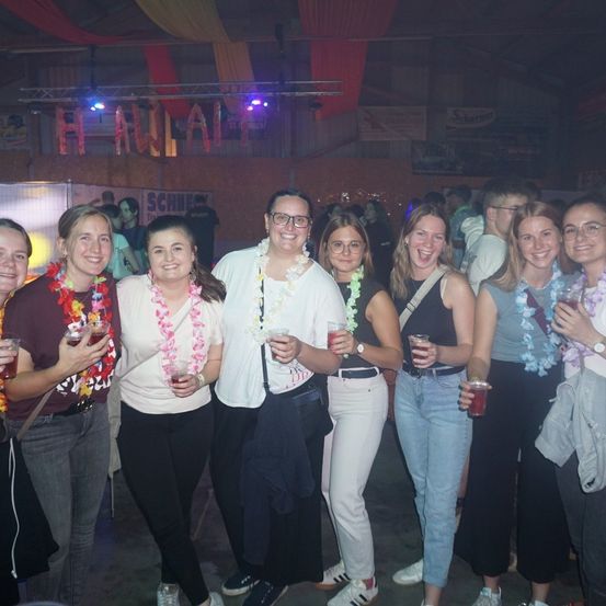 Eine Gruppe von Frauen mit Blumenketten und Getränken in der Hand posiert für ein Foto in einem schwach beleuchteten Raum mit einem Schild, das 'Hawaii' steht.
