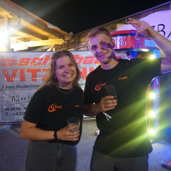 Ein Mann und eine Frau stehen bei Nacht, lächeln und halten Getränke. Sie tragen passende schwarze T-Shirts mit orangefarbenen Logos. Hinter ihnen ist ein Banner mit rotem Text und eine rote Box mit grünen Lichtern zu sehen.