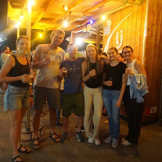 Eine Gruppe von fünf Personen, vier Frauen und ein Mann, posiert für ein Foto mit Biergläsern in der Hand. Sie stehen vor einer Holzwand mit gelben Lichtern.