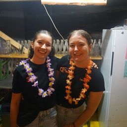 Zwei Frauen in schwarzen T-Shirts mit Blumenketten, die lächelnd für ein Foto in einem Raum mit einem Holzregal und einem Kühlschrank im Hintergrund posieren.