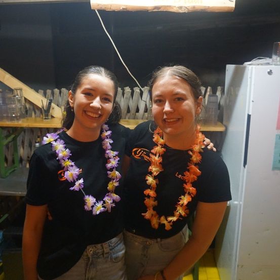 Zwei Frauen in schwarzen T-Shirts mit Blumenketten, die lächelnd für ein Foto in einem Raum mit einem Holzregal und einem Kühlschrank im Hintergrund posieren.