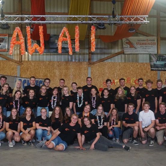 Eine Gruppe von Menschen in passenden schwarzen T-Shirts mit orangefarbenen Leis um den Hals posiert für ein Foto in einem Raum mit einem großen 'AWAII'-Schild über ihnen.