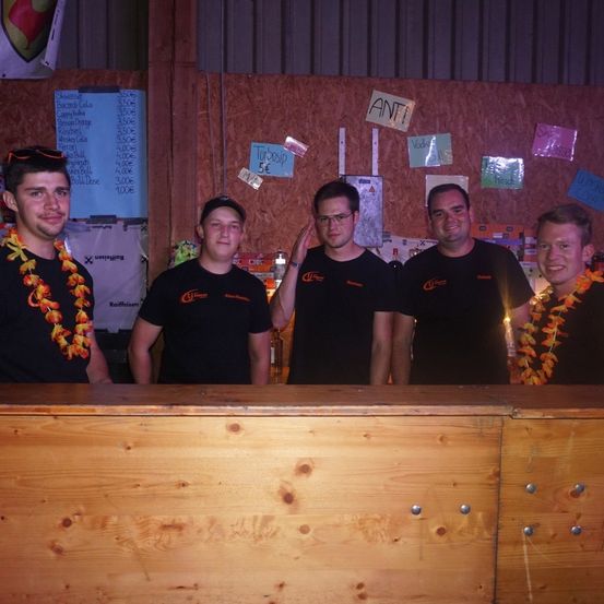 Fünf Männer in schwarzen T-Shirts mit orangefarbenen Girlanden stehen hinter einer Holzbar und lächeln für die Kamera. Hinter ihnen ist eine Wand mit verschiedenen Plakaten und Preisschildern zu sehen.