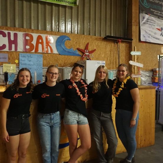 Fünf Frauen stehen zusammen vor einer Theke, tragen passende schwarze T-Shirts und Blumenkränze. Sie scheinen für ein Foto zu lächeln, mit einem Banner, das 'Beach Bar' über ihnen steht.
