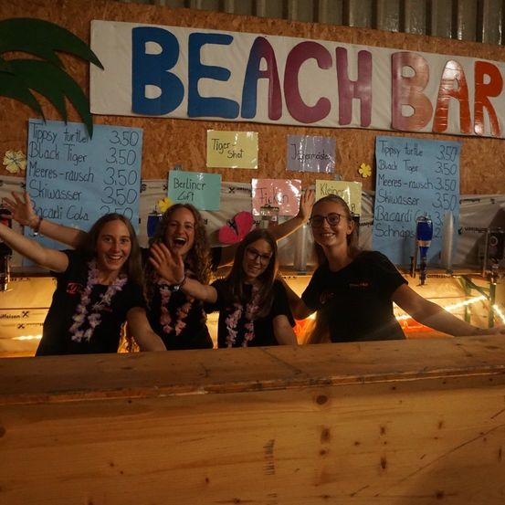 Vier Frauen posieren für ein Foto an einer Bar mit einem Schild, das Beach Bar steht. Sie tragen Blumenketten und lächeln. Die Bar hat eine Holztheke und ein Menübrett mit Preisen.
