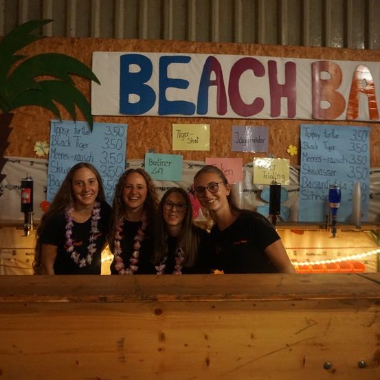 Vier Frauen stehen hinter einer Bartheke und lächeln für ein Foto. Sie tragen schwarze T-Shirts und Blumenketten. Hinter ihnen zeigt eine Wand ein Schild mit der Aufschrift 'Beach Bar' und verschiedene Preisschilder.