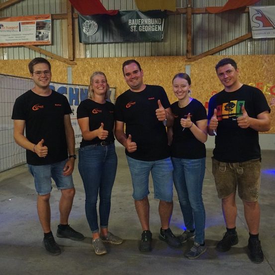 Fünf Personen in identischen schwarzen T-Shirts posieren für ein Foto und zeigen Daumen hoch. Sie stehen in einem Raum mit einem Banner, das 'Bauernbund St. Georgen' besagt.