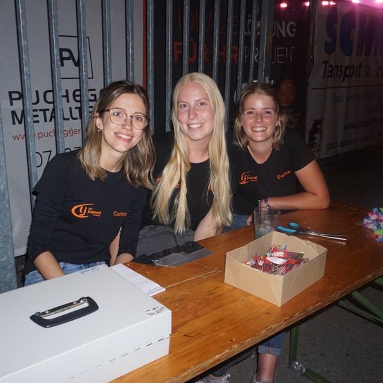 Drei Frauen in schwarzen T-Shirts mit orangefarbenen Logos sitzen an einem Tisch und lächeln für die Kamera. Eine Schachtel mit Süßigkeiten und Scheren sind auf dem Tisch. Dahinter befindet sich ein Metallzaun mit einem Banner.