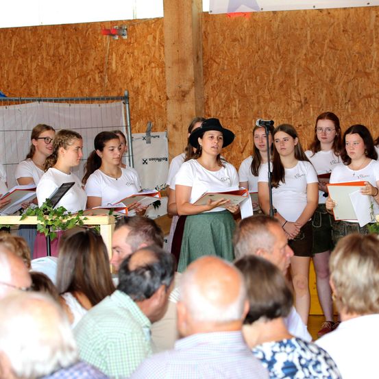 Ein Chor junger Frauen in weißen Shirts und Röcken steht auf der Bühne und hält Notenblätter in der Hand, vor ihnen ein Publikum.