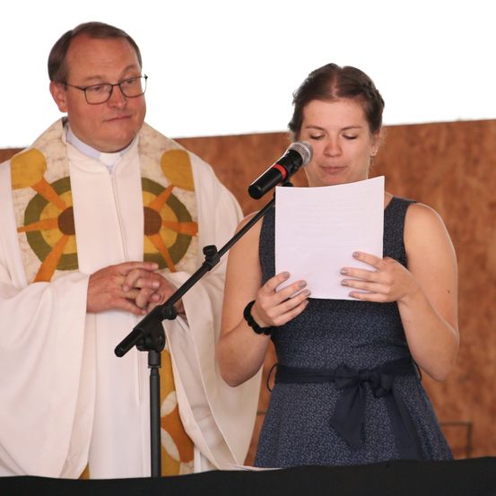 Eine Frau liest von einem Papierstück an einem Rednerpult, mit einem Priester hinter ihr.