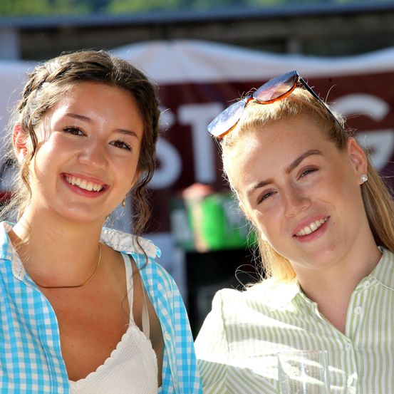 Zwei Frauen lächeln und posieren für ein Foto, eine mit Sonnenbrille auf dem Kopf und die andere mit einer Halskette.