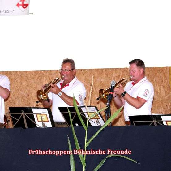 Drei Männer in weißen Hemden spielen Trompeten auf der Bühne, mit einem Mikrofon und Notenständern vor ihnen. Der Hintergrund zeigt eine Holzwand und ein Banner mit Text.