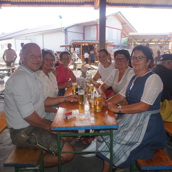 Eine Gruppe von Menschen sitzt an einem Tisch in einem Biergarten, lächelt und genießt ihre Getränke. Der Tisch ist mit Biergläsern und einer Schachtel Zigaretten gefüllt.