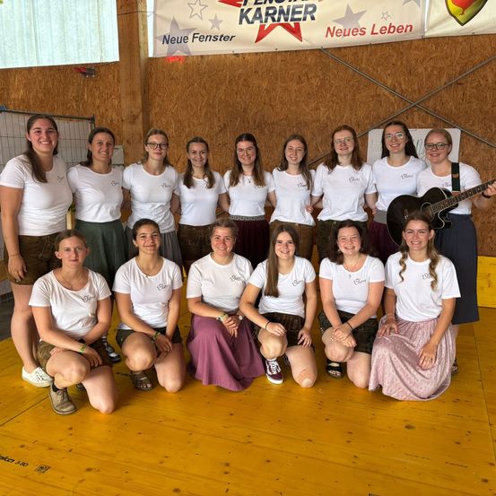 Eine Gruppe von Frauen in weißen Shirts und Röcken, die für ein Foto in einem Raum mit Holzboden und einem Banner mit der Aufschrift 'Neue Fenster Neues Leben' posieren.
