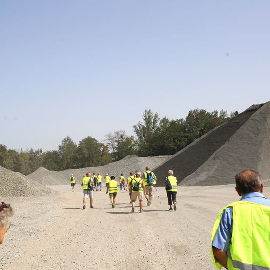 Eine Gruppe von Menschen in neongelben Westen und mit Rucksäcken geht auf einem Schotterweg, mit großen Haufen von Schotter auf beiden Seiten.