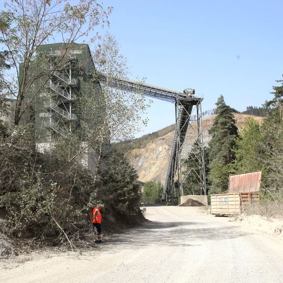 Eine Person in einer orangefarbenen Weste geht einen Feldweg entlang, mit einer großen Industrieanlage im Hintergrund.