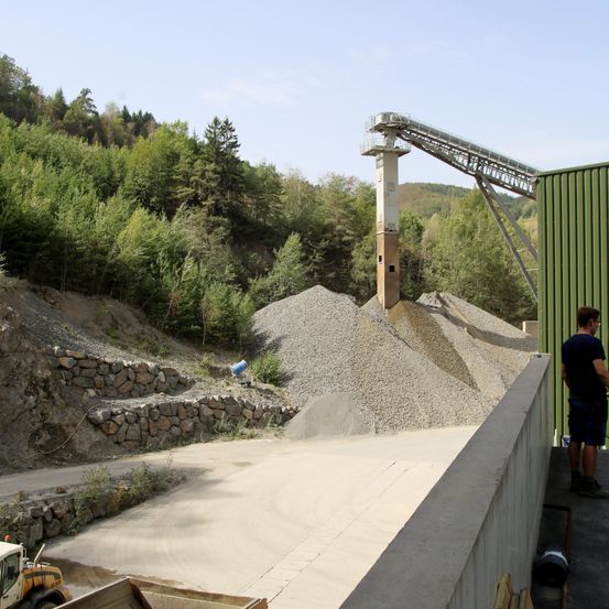Eine Baustelle mit einem großen Kieshaufen und einem Förderbandsystem. Ein Mann steht auf einer Betonplattform in der Nähe einer grünen Wand.