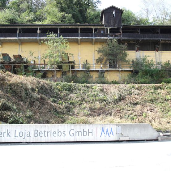Ein altes gelbes Industriegebäude mit schwarzem Dach und einigen rostigen Geräten auf dem Boden. Das Schild steht 'Werk Loja Betriebs GmbH'.