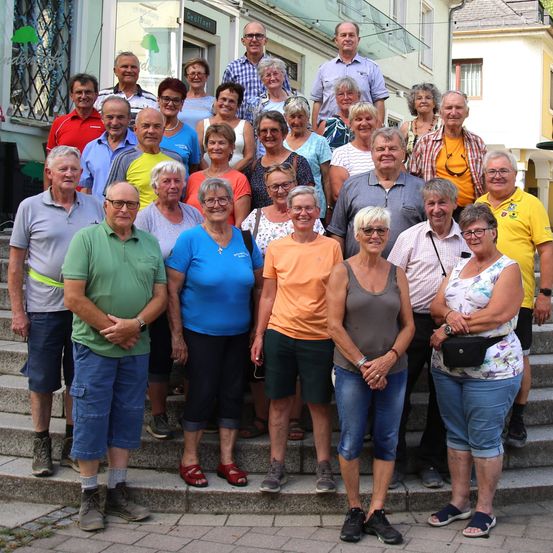 Eine Gruppe älterer Menschen posiert für ein Foto auf einer Freitreppe. Sie tragen Freizeitkleidung und einige tragen Brillen. Hinter ihnen steht ein Gebäude mit Glasfenstern und einem Metallzaun.