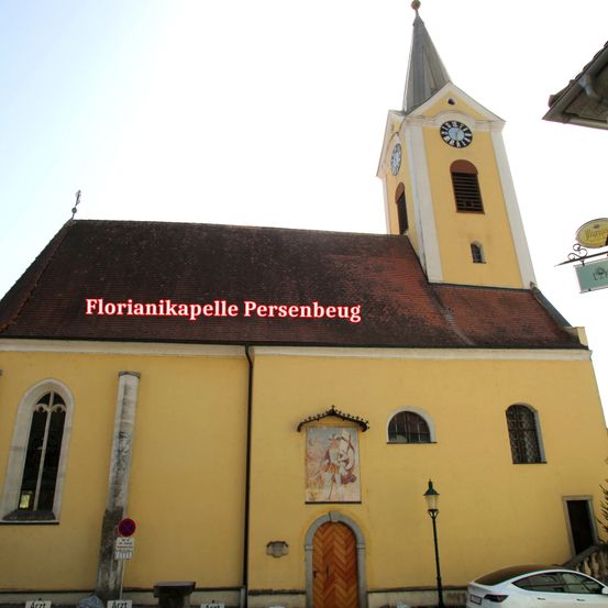 Eine gelbe Kirche mit einem Turm und einem Glockenturm, namens Florianikapelle Persenbeug, mit einem Schild an der Seite und einem weißen Auto, das davor geparkt ist.