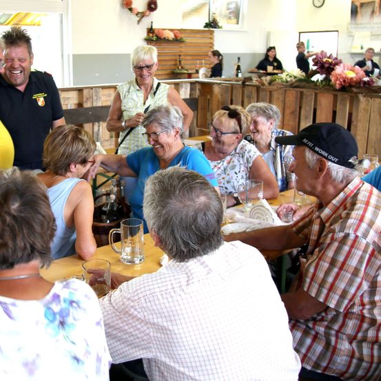 Eine Gruppe älterer Erwachsener versammelt sich in einem Restaurant, lächelt und unterhält sich. Einige tragen Brillen und Freizeitkleidung. Auf dem Tisch stehen Gläser, Teller und Blumen. Ein Mann in einem schwarzen Shirt steht hinter ihnen.