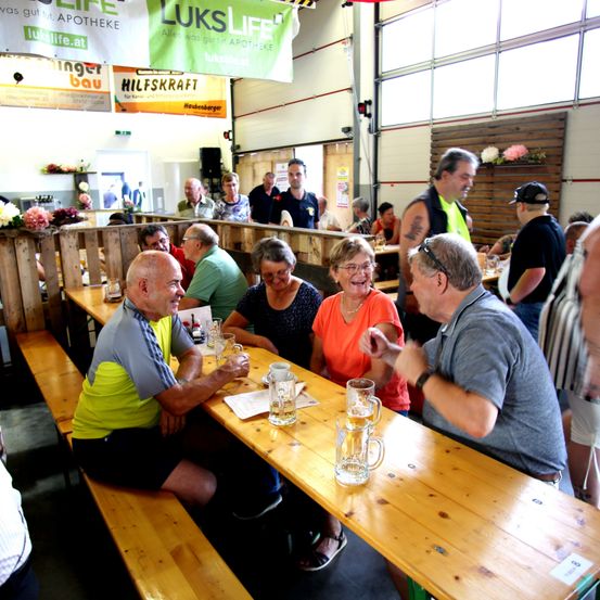 Eine Gruppe von Menschen sitzt an einem langen Holztisch in einem Restaurant, lächelt und lacht. Es gibt Gläser Bier und Tassen auf dem Tisch. Das Restaurant hat Holzwände und Banner hängen an der Decke.