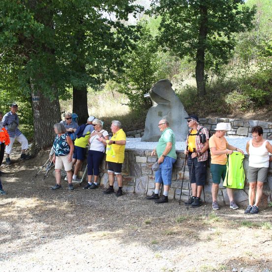 Eine Gruppe von Wanderern in Freizeitkleidung, einige mit Hüten, stehen um ein Steinmonument in einem bewaldeten Gebiet. Bäume und Gras umgeben die Szene.