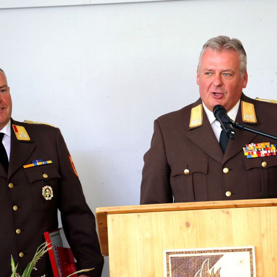 Zwei Männer in Militäruniformen, einer spricht an einem Podium mit Mikrofon, der andere steht in der Nähe.