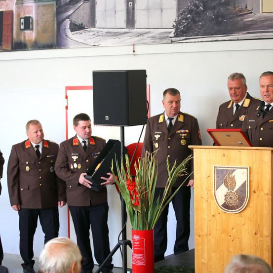 Fünf Männer in Militäruniformen stehen an einem Podium mit Mikrofon und Laptop, einer hält eine Schachtel und Blumen.