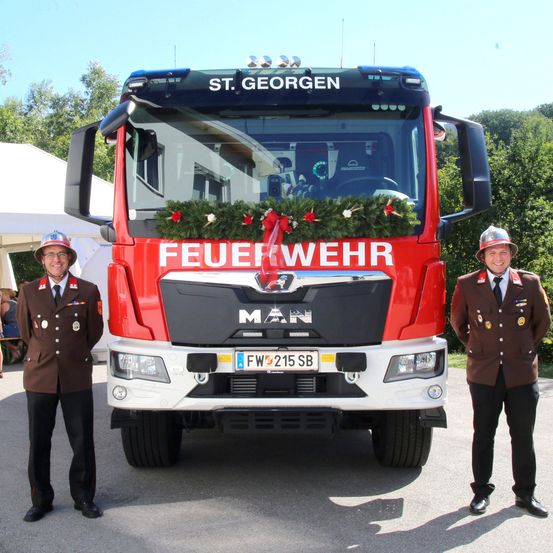 Zwei Feuerwehrleute in Uniform stehen vor einem roten Feuerwehrauto mit 'St. Georg' und 'Feuerwehr' darauf. Das Auto hat eine Girlande vorne.