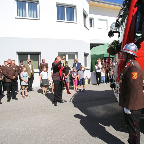 Ein Mann in einem roten Gewand und Hut steht vor einem roten Feuerwehrauto, während mehrere uniformierte Personen und andere zuschauen.