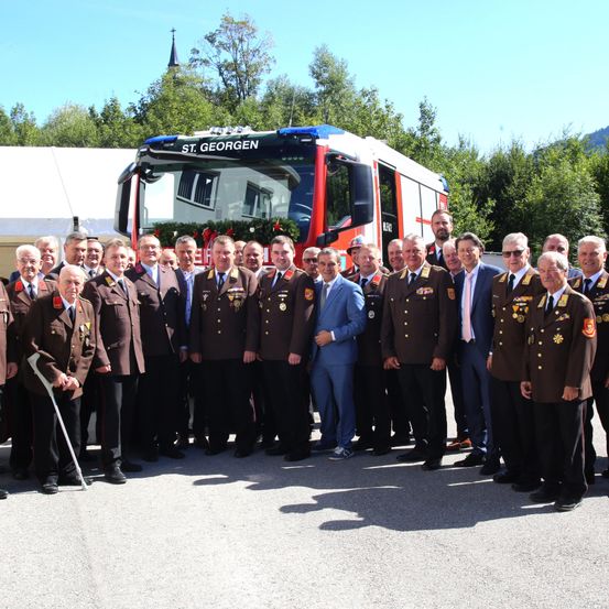 Eine Gruppe von Männern in Militäruniformen und Anzügen steht vor einem Feuerwehrauto mit der Aufschrift 'St. Georgen'. Sie posieren für ein Foto.