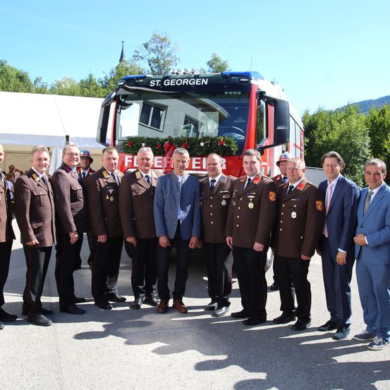 Eine Gruppe von Feuerwehrleuten und Beamten steht vor einem Feuerwehrwagen, wahrscheinlich für ein Foto. Sie tragen alle Uniformen und einige haben Medaillen.